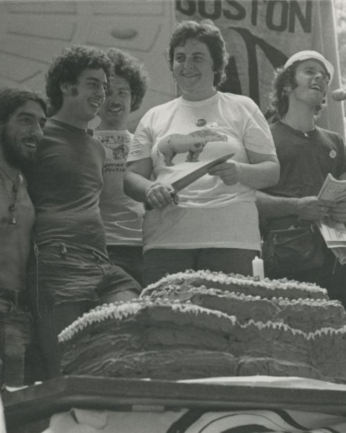 Photo shows the cake cutting a little later during the Pride rally on the Common. L-R: unidentified child, Ron Arruda, Jared Goldfine, John Kyper, Ellen B. Davis (with knife), Bernie Toale and Jim Saslow.